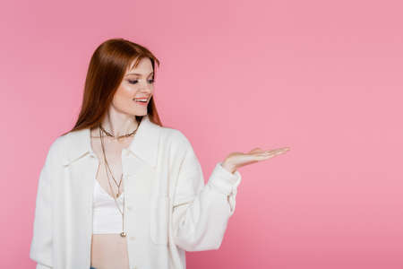 Smiling red haired woman in jacket pointing with hand isolated on pinkの写真素材