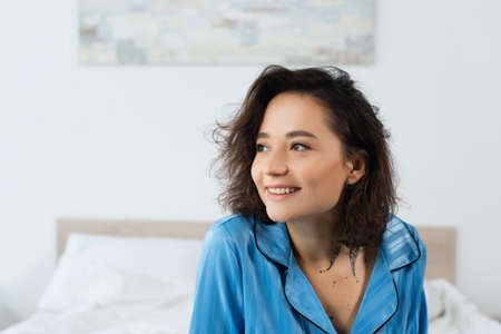 happy and tattooed woman in blue pajamas looking away at home