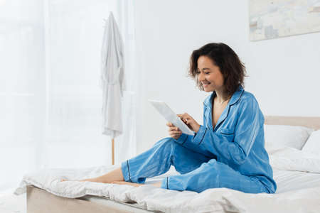 cheerful young woman in blue pajamas using digital tablet in bedroomの写真素材