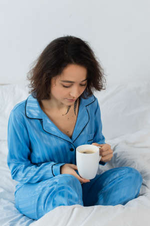 young woman in blue pajamas holding cup of coffee in bedroomの写真素材