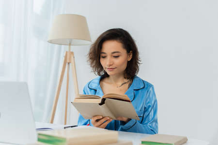 tattooed young woman reading book near laptop on desk