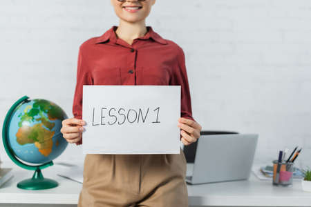 cropped view of smiling woman holding paper with lesson one lettering near globe and laptop on deskの写真素材