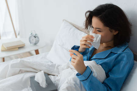 sick woman sneezing in tissue and looking at electronic thermometer