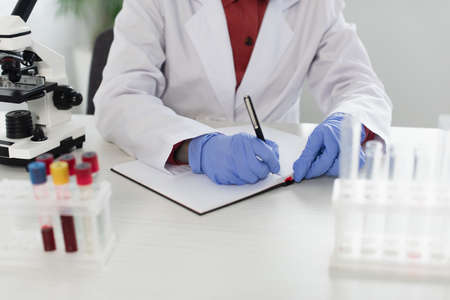 cropped view of scientist in latex gloves writing in notebook near test tubes and microscope