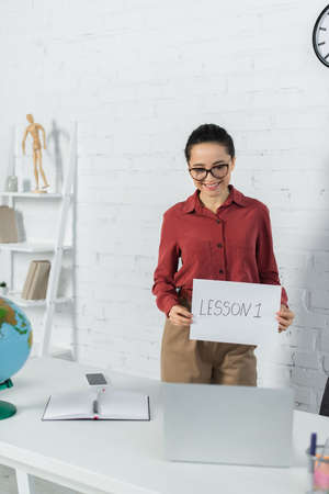 smiling teacher in eyeglasses holding paper with lesson one lettering and looking at laptopの写真素材