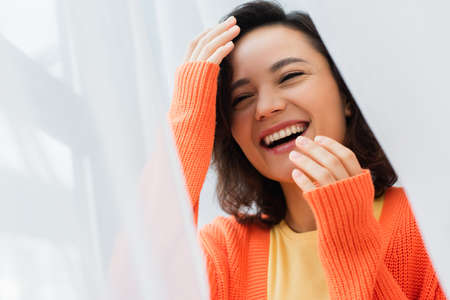 portrait of cheerful young woman laughing near white curtain