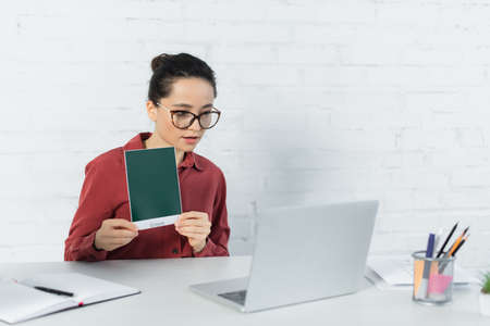 young teacher in eyeglasses holding card with green lettering and looking at laptop on desk