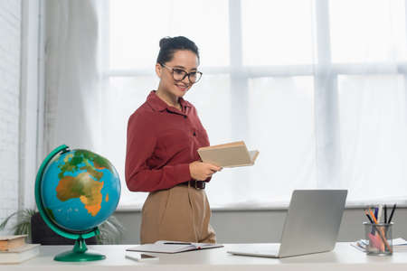 cheerful teacher in eyeglasses holding book and looking at laptop near smartphone on desk