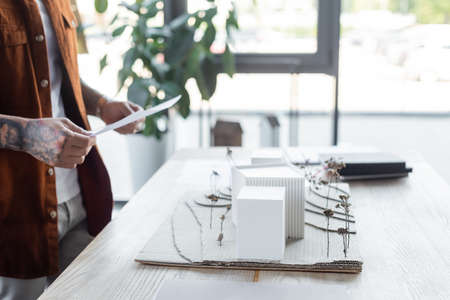 partial view of tattooed engineer holding document near blurred house models on deskの写真素材