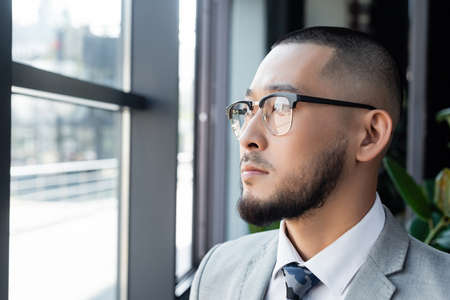 bearded asian businessman in eyeglasses looking away near window in office