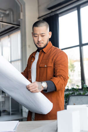 asian architect looking at blurred house models while holding blueprint in officeの写真素材