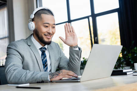 smiling asian businessman in headphones waving hand during video call on laptop in officeの写真素材