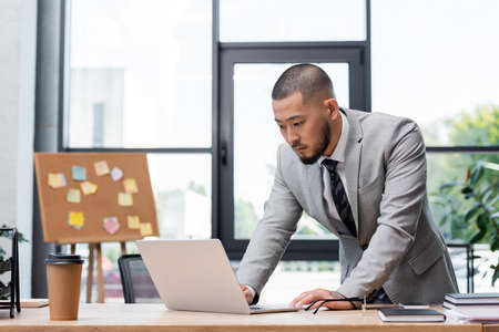 bearded asian businessman standing near work desk and looking at laptop in officeの写真素材