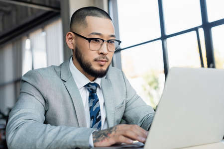 asian businessman in eyeglasses typing on blurred laptop in office