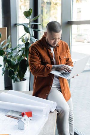 concentrated asian architect typing on laptop while sitting on desk near blueprints and house models