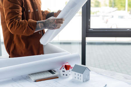 cropped view of tattooed architect holding blueprint while sitting on desk near calculator and house...