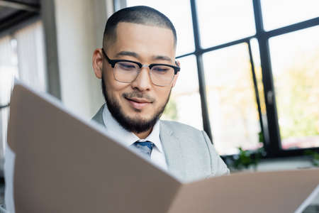 smiling asian businessman looking at documents in blurred folder in office