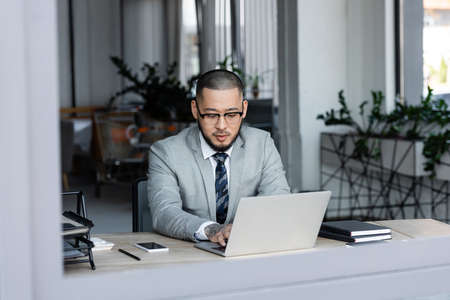 asian businessman in eyeglasses working on laptop in office on blurred foregroundの写真素材