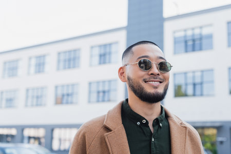cheerful asian man in stylish sunglasses smiling near blurred building on urban streetの写真素材