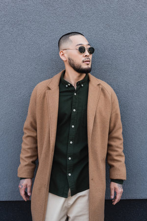 bearded asian man in autumn coat and sunglasses standing near grey wallの写真素材