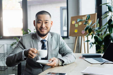 happy asian businessman with cup of coffee looking at camera while sitting at workplace