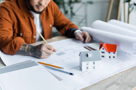 cropped view of blurred architect drawing blueprint near house models on deskの写真素材