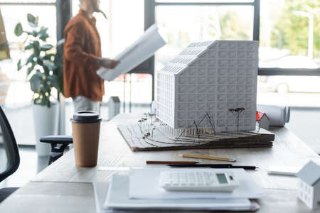 selective focus of house model and paper cup on desk near architect standing with paper on blurred backgroundの写真素材