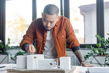 tattooed asian architect measuring house models with pencil while working in office