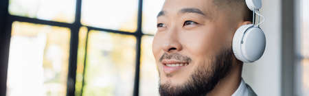 smiling asian businessman in wireless headphones looking away in office, banner