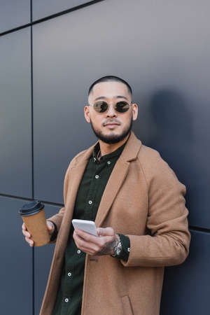 asian man in trendy autumn outfit holding takeaway drink and mobile phone near grey wallの写真素材