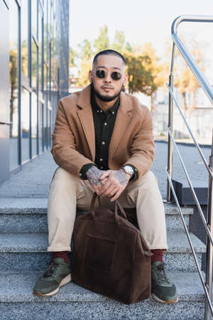 stylish asian man in beige coat and sunglasses sitting on stairs with leather briefcase