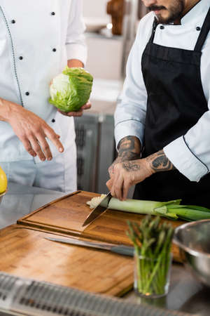 Cropped view of chef cutting leek near colleague with cabbage in kitchenの写真素材