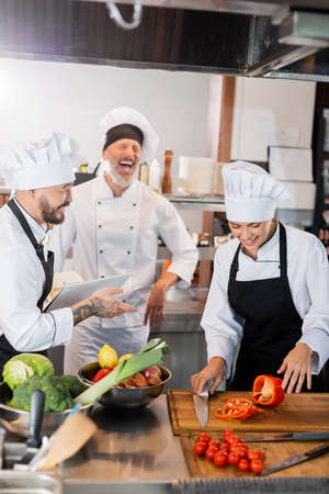 Asian chef holding cookbook near laughing colleagues and fresh food in kitchenの写真素材