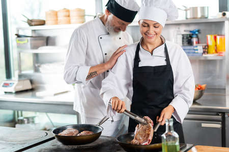Chef standing near smiling colleague roasting meat in kitchen