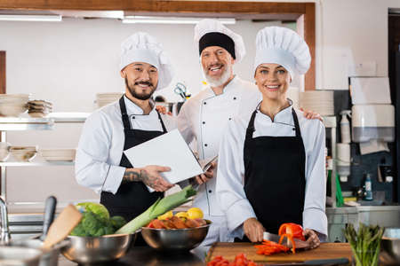 Asian chef holding cookbook near smiling colleagues and vegetables in kitchenの写真素材