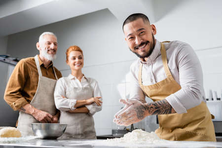 Positive asian chef in apron standing near flour and blurred colleagues in kitchenの写真素材