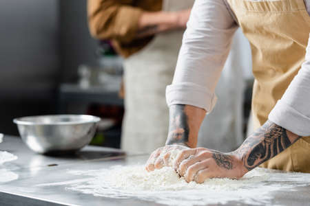 Cropped view of tattooed chef making dough near blurred colleague in kitchen