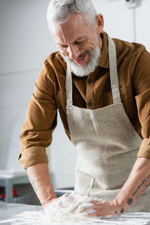 Smiling mature chef in apron making dough in kitchenの写真素材