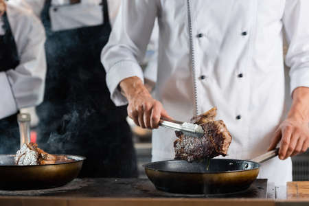 Cropped view of chef holding meat in tongs near frying pans and colleagues in kitchen