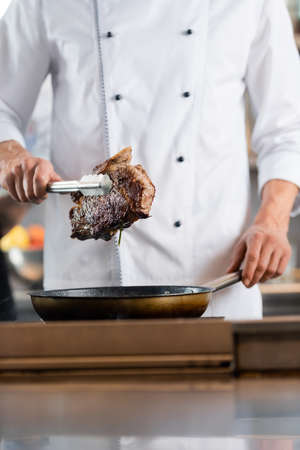 Cropped view of chef holding roasted meat in tongs near frying pan