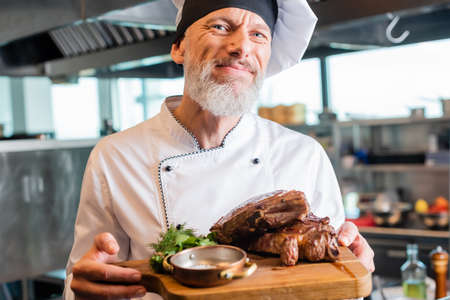 Mature chef smiling at camera while holding roasted meat on cutting board in kitchenの写真素材