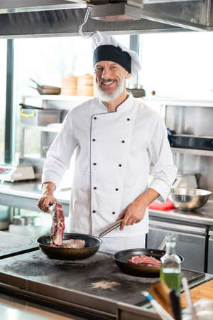 Smiling chef in uniform cooking meat on cooktop in restaurant kitchenの写真素材