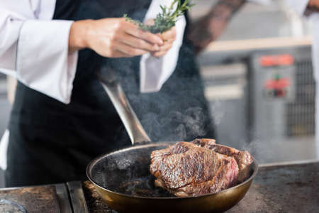 Roasted meat on frying pan near blurred chef holding rosemary in kitchenの写真素材
