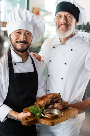 Positive multiethnic chefs holding cooked meat on cutting board in kitchenの写真素材