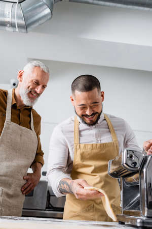 Cheerful asian chef holding dough near pasta maker machine and colleague in kitchenの写真素材