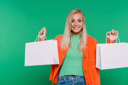 cheerful blonde woman with shopping bags smiling at camera isolated on greenの写真素材