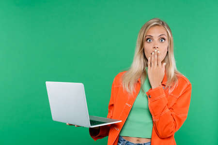 nervous blonde woman covering mouth with hand while holding laptop isolated on greenの写真素材