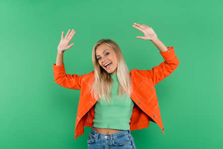 excited blonde woman in jeans and orange jacket waving hands isolated on green