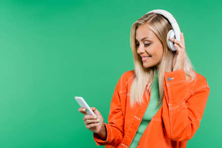 smiling blonde woman looking at mobile phone while listening music in headphones isolated on greenの写真素材