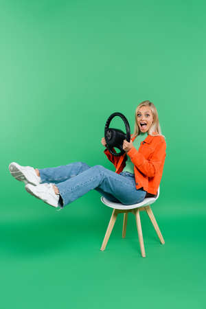 astonished woman with steering wheel looking at camera while sitting on chair with raised legs on greenの写真素材